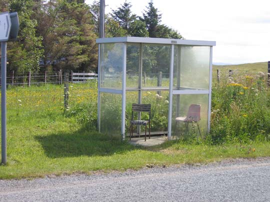 An enclosed bus shelter along a country lane with two chairs that someone has put there out of necessity